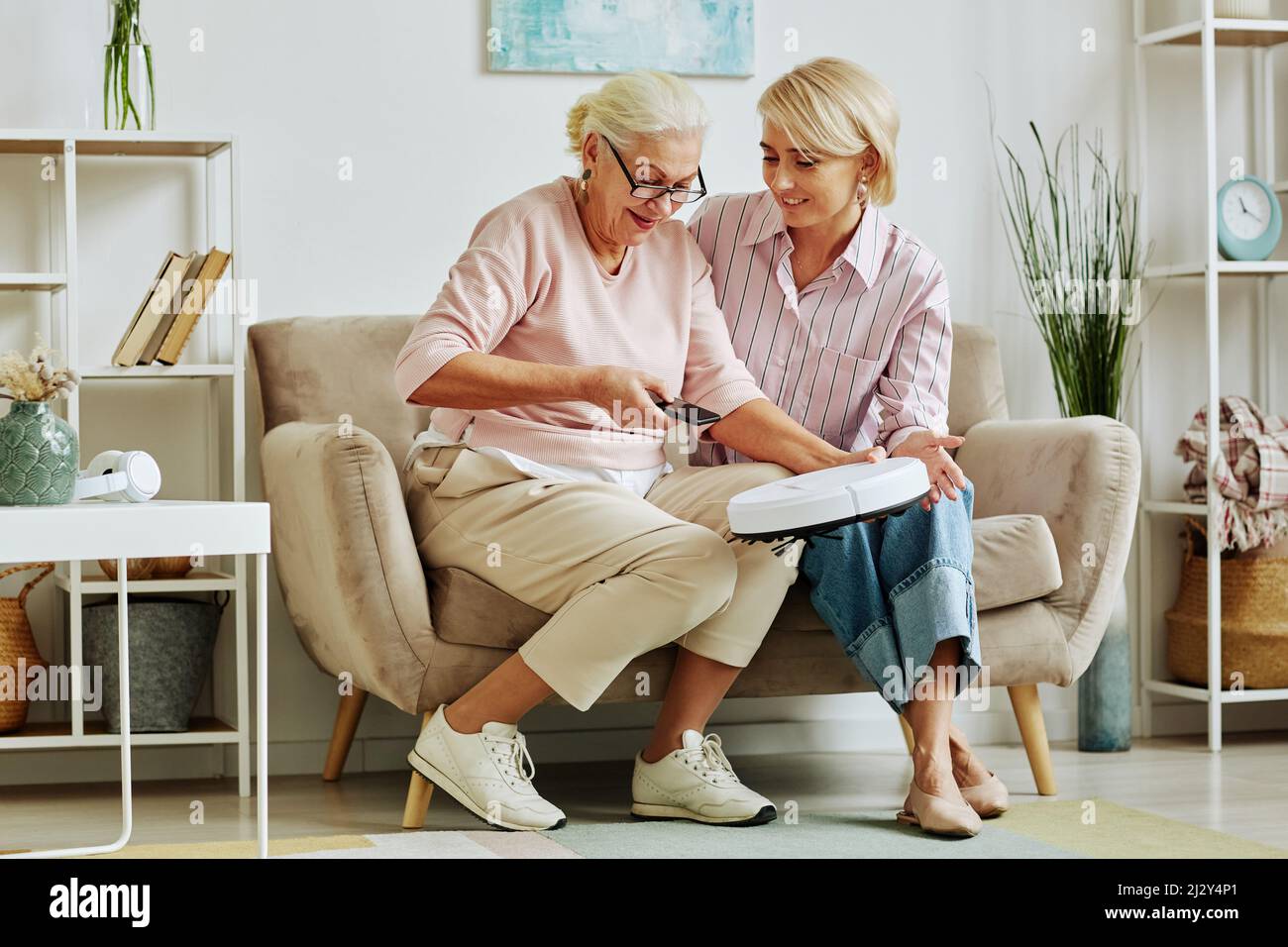 Ritratto completo di donna anziana sorridente che collega l'aspirapolvere robot allo smartphone durante la configurazione di dispositivi domestici intelligenti con la nuora adulta Foto Stock
