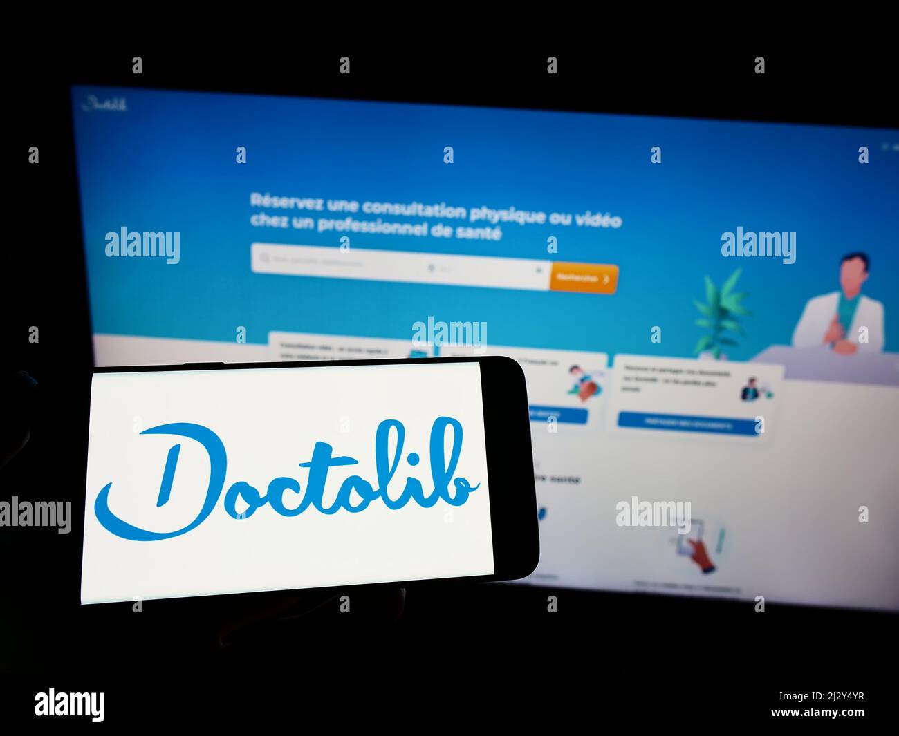 Persona che tiene uno smartphone con il logo della società francese di assistenza sanitaria Doctolib SAS sullo schermo di fronte al sito web. Mettere a fuoco sul display del telefono. Foto Stock