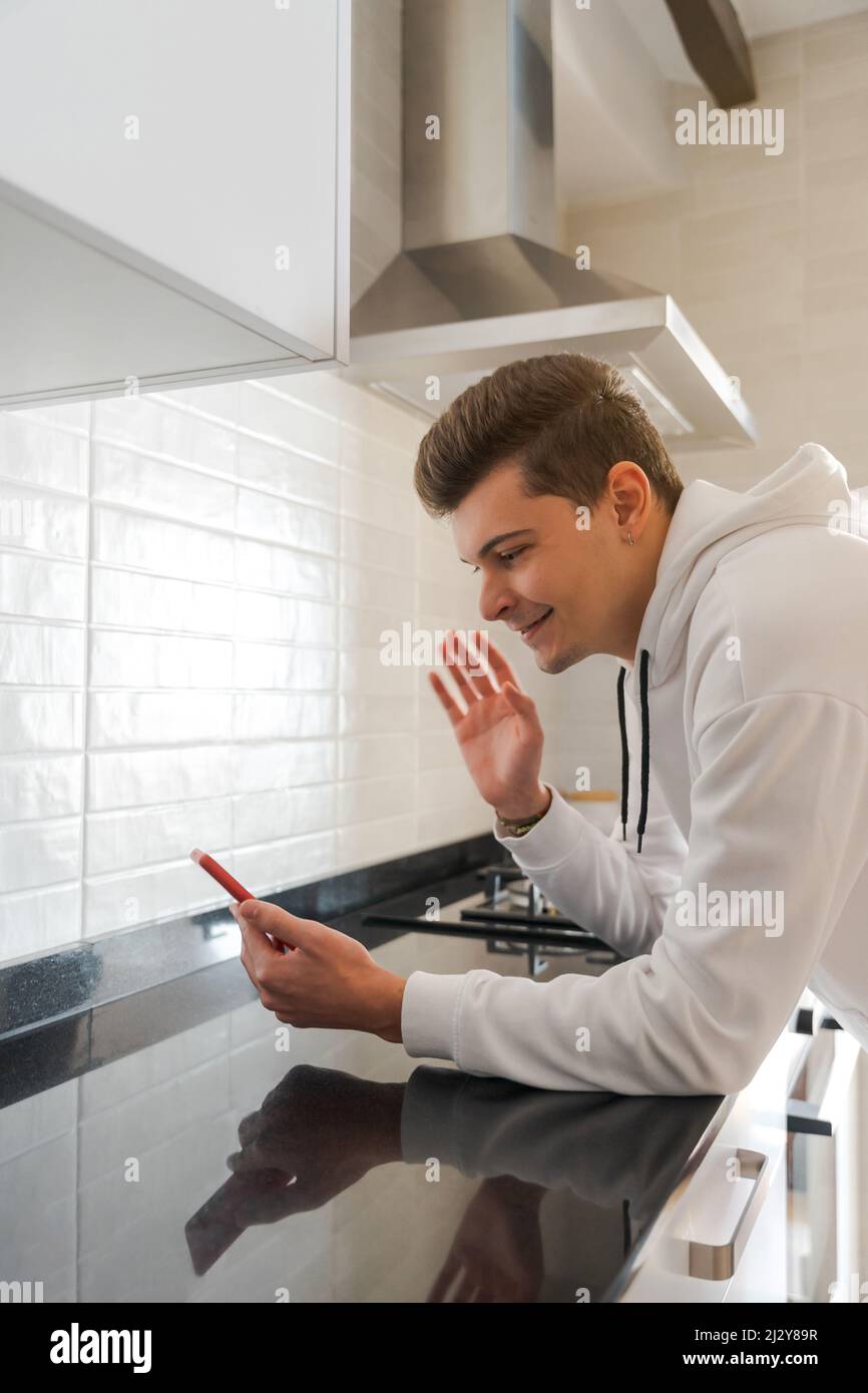 Il giovane uomo caucasico sorrise appoggiandosi sul banco della cucina. Sta agitando e facendo una videochiamata sul suo cellulare. Si accende la luce naturale. Lui io Foto Stock