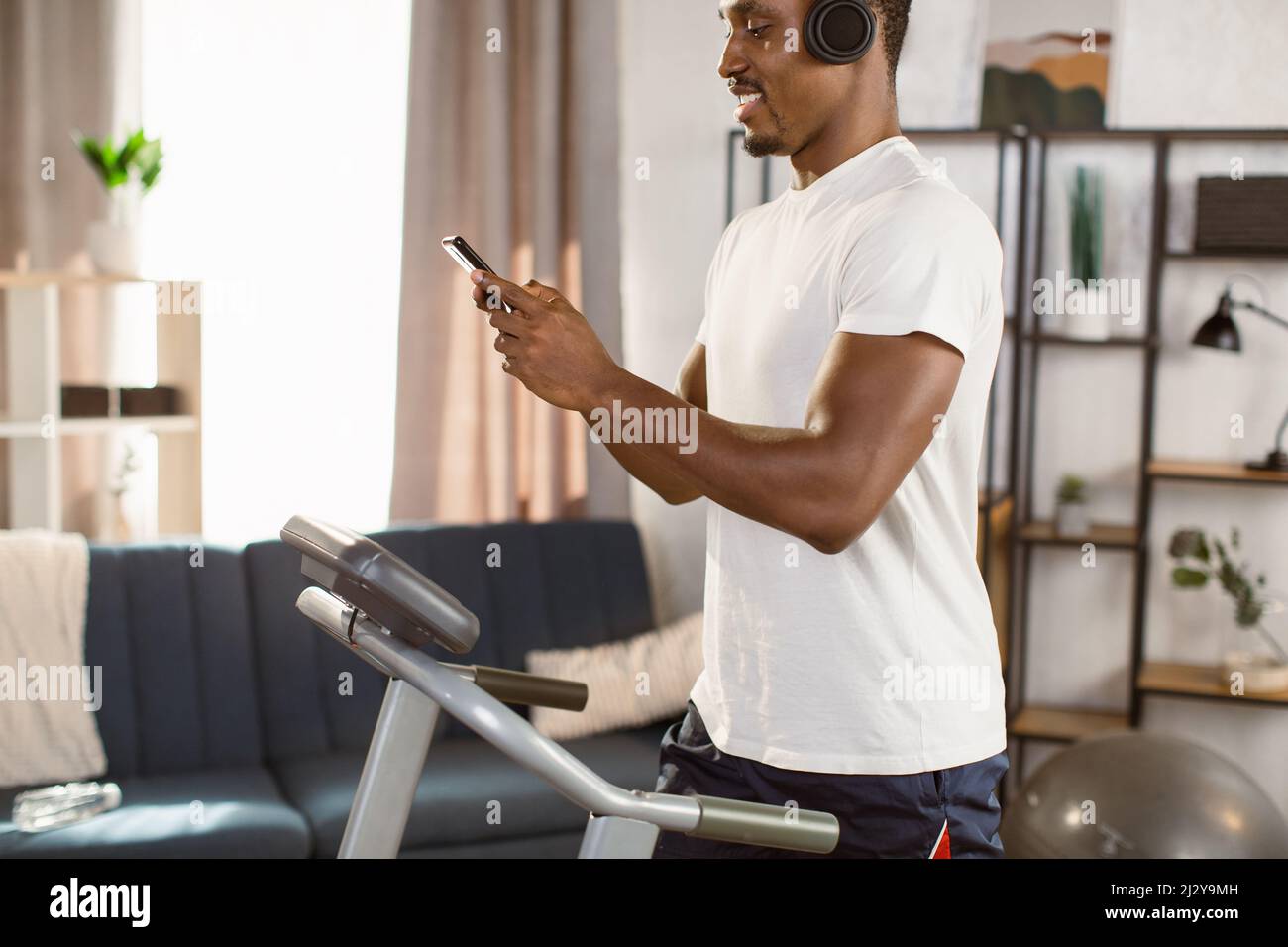 Immagine ritagliata dell'uomo africano che corre sul tapis roulant con il telefono in mano e le cuffie in un moderno appartamento luminoso. Concetto di stile di vita sano. Youn Foto Stock