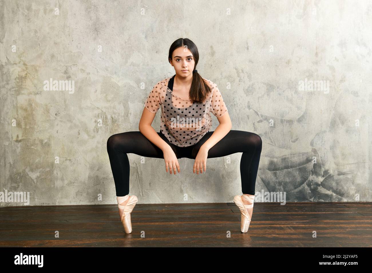 Vista frontale della giovane ballerina in posa en punto bilanciamento sulle punte delle dita dei piedi in speciali scarpe balletto contro una parete grunge grigio studio in cerca di quie Foto Stock