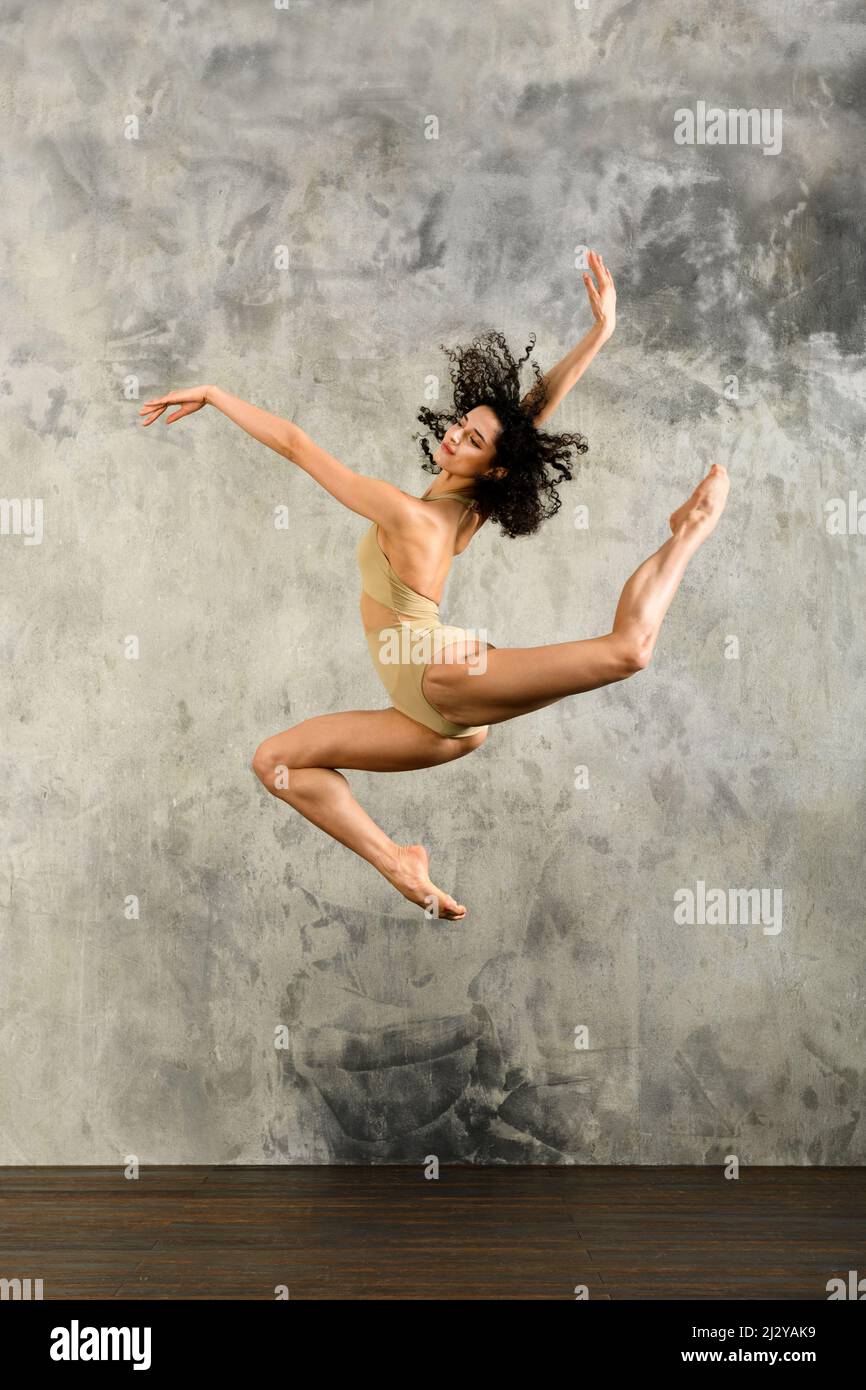 Vista laterale del corpo della ballerina attiva a piedi nudi in bodysuit esecuzione salto con braccia sollevate in studio luce durante le prove Foto Stock
