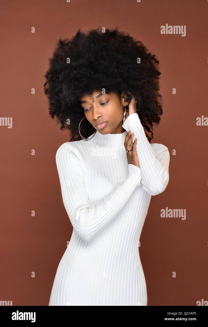 Slender calma signora dominicana con acconciatura Afro in abito bianco che tocca i capelli e guarda in giù su sfondo marrone Foto Stock