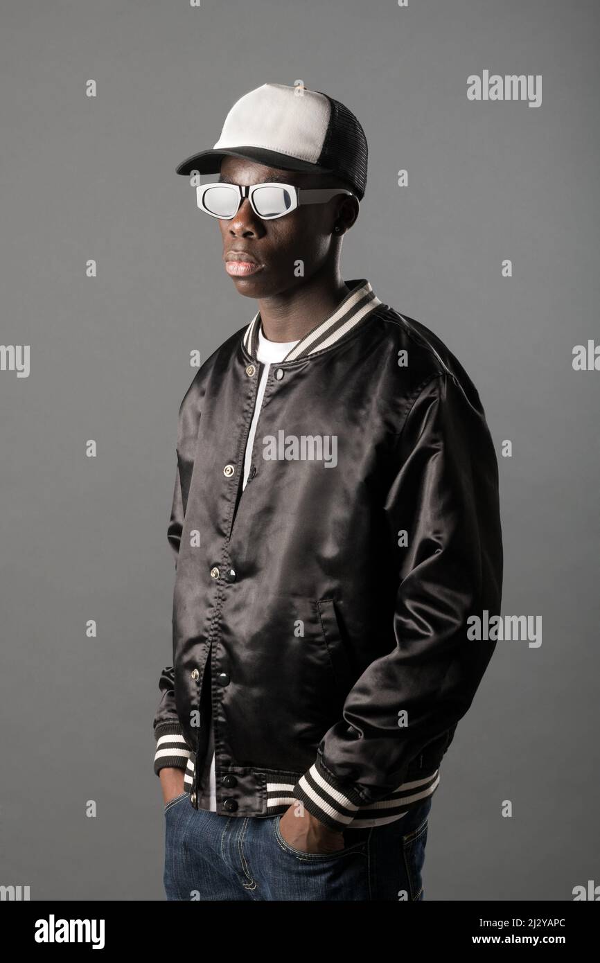 Fidato rapper maschile afroamericano in berretto e occhiali da sole in piedi con le mani su tasche su sfondo grigio in studio chiaro Foto Stock