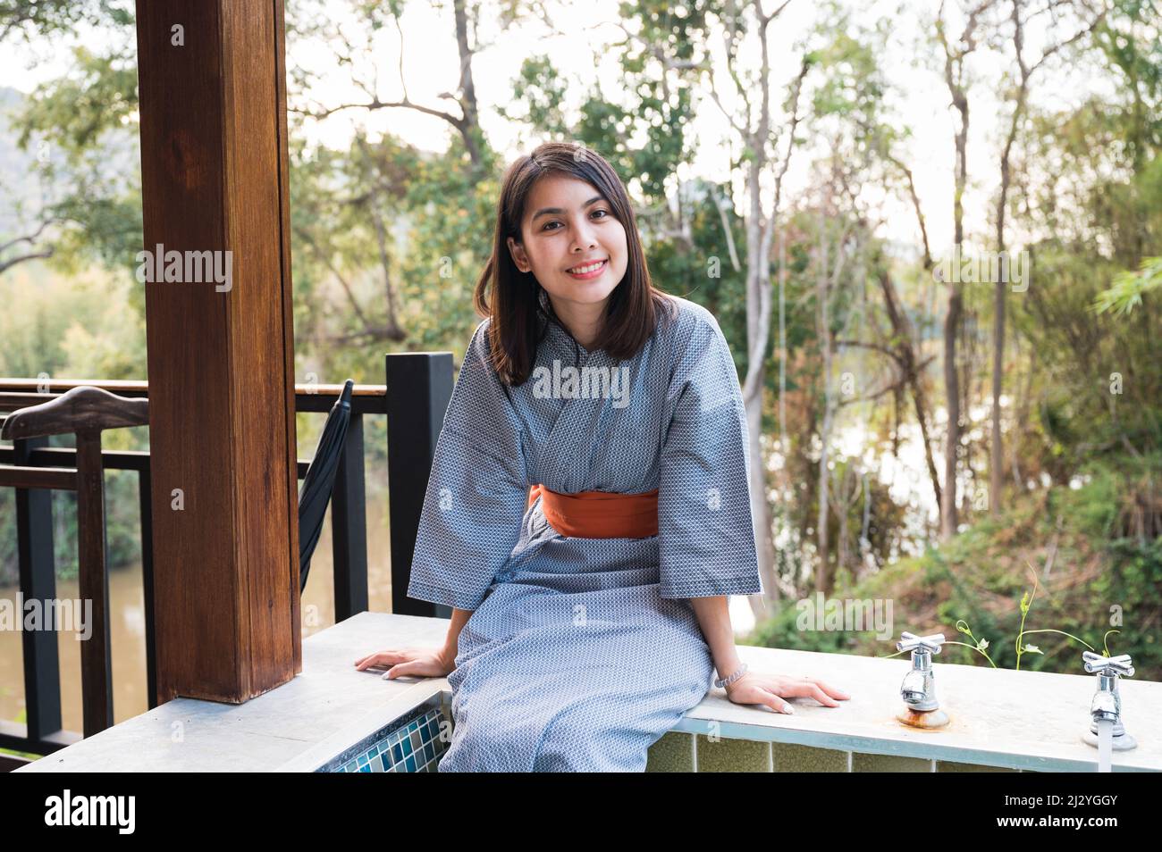 Allegra giovane donna asiatica in accappatoio seduta sulla piscina termale calda naturale tra la natura del resort Foto Stock