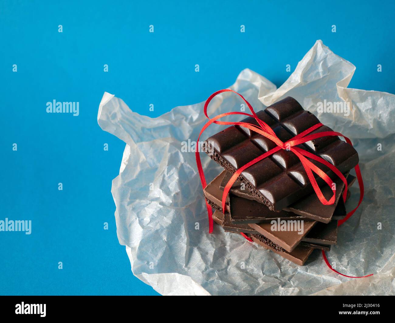 Pezzi di cioccolato con un nastro rosso su sfondo blu, spazio copia. Concetto di sorpresa, dolce dono, amore e romanticismo Foto Stock