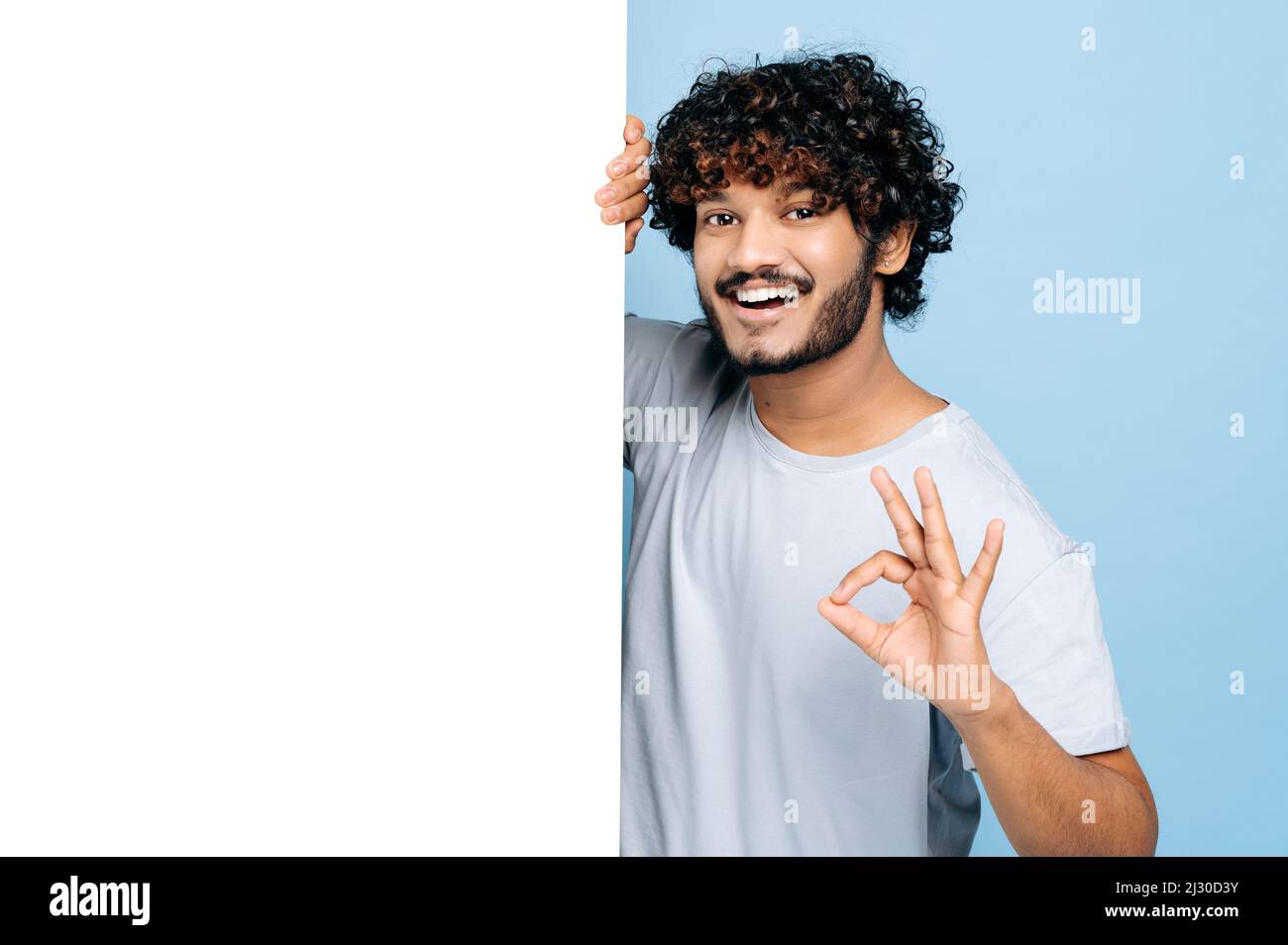 Ragazzo indiano o arabo felice, in T-shirt casual, sbirciando fuori da dietro la lavagna bianca della pubblicità, mostrando spazio vuoto della copia per il vostro testo o disegno, mostra IL gesto GIUSTO, sfondo blu isolato Foto Stock