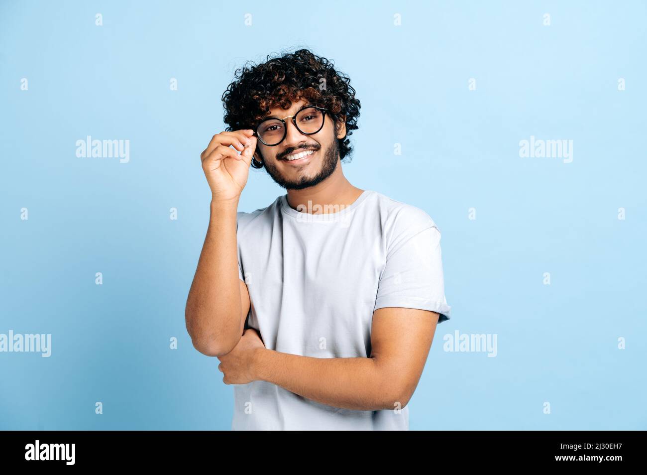 Attraente bel ragazzo positivo indiano o arabo trendy ricci-capelli con occhiali, freelancer o designer creativo, si leva su uno sfondo blu isolato, guarda la fotocamera, sorride amichevole Foto Stock