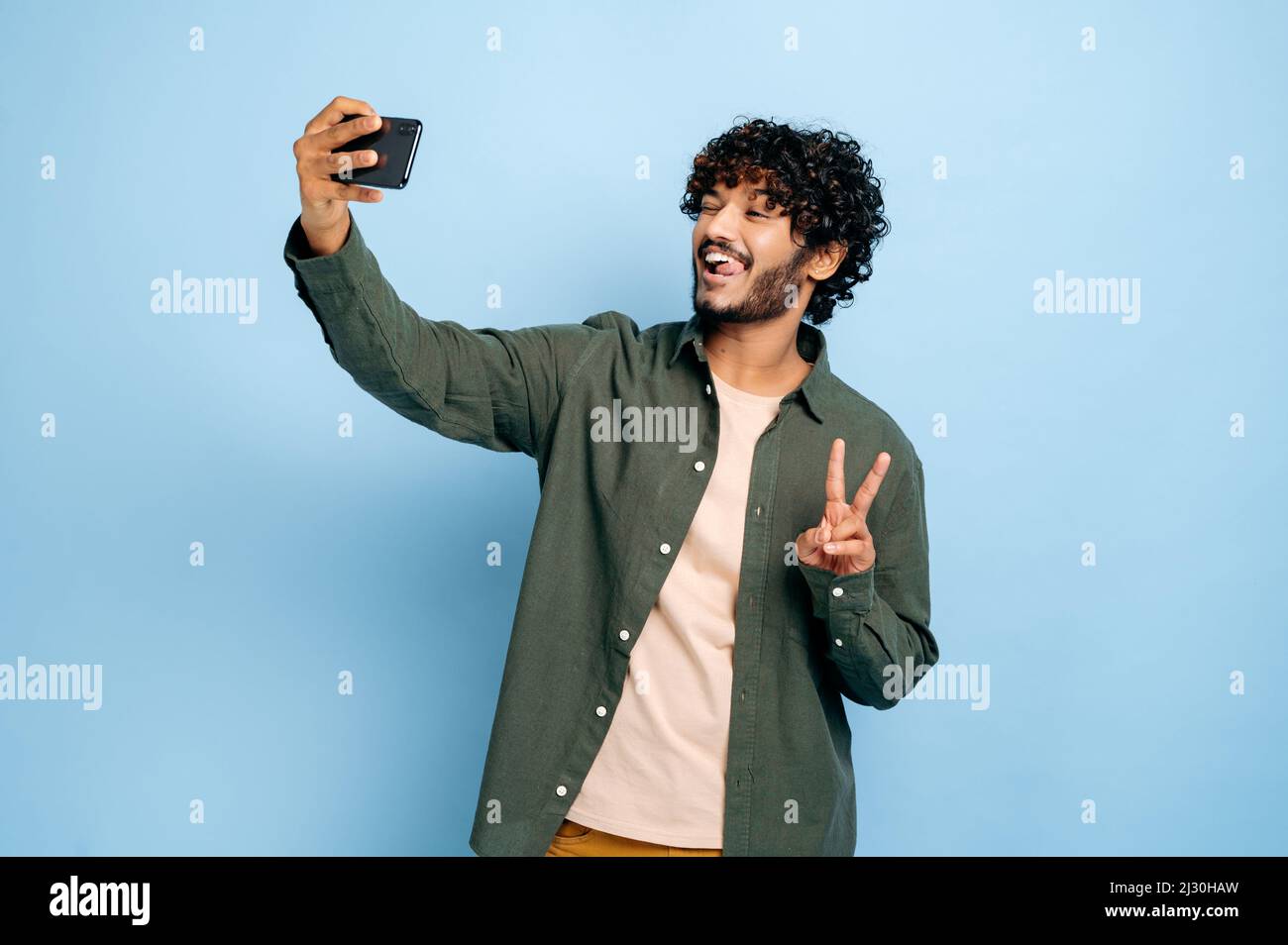 Felice positivo indiano o arabo eccitato ragazzo, in abbigliamento casual elegante, facendo selfie girato sul cellulare e sorride nella fotocamera, mostra simbolo di pace, posando su isolato sfondo blu Foto Stock