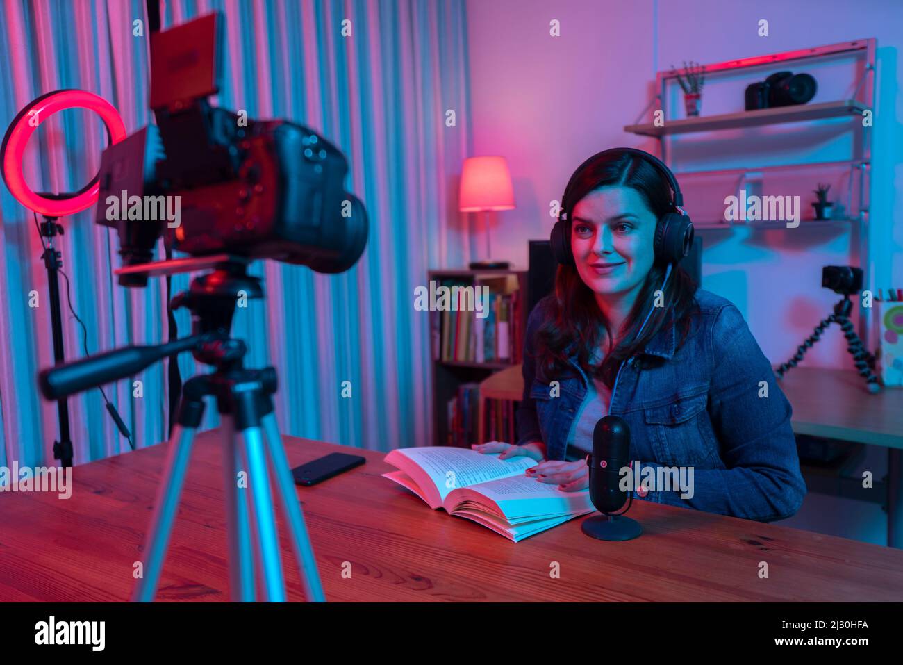 Bella donna ispanica di fronte a una videocamera che registra un blog nel suo studio con luci rosse e blu all'interno della sua casa. Trasmissione della comunicazione Foto Stock