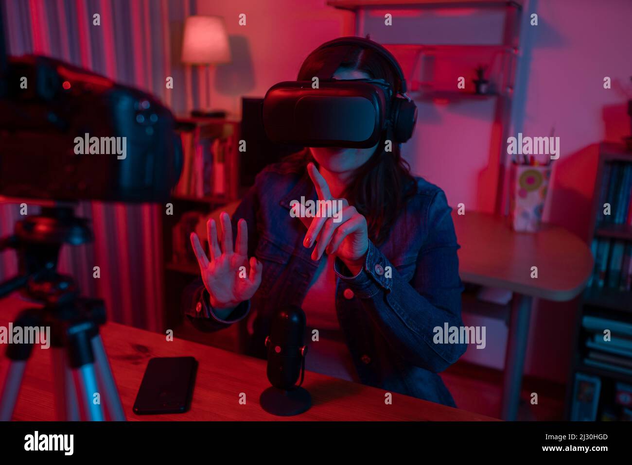 Bella donna ispanica di fronte a una videocamera che registra un blog nel suo studio con luci rosse e blu all'interno della sua casa. Trasmissione con VR gla Foto Stock