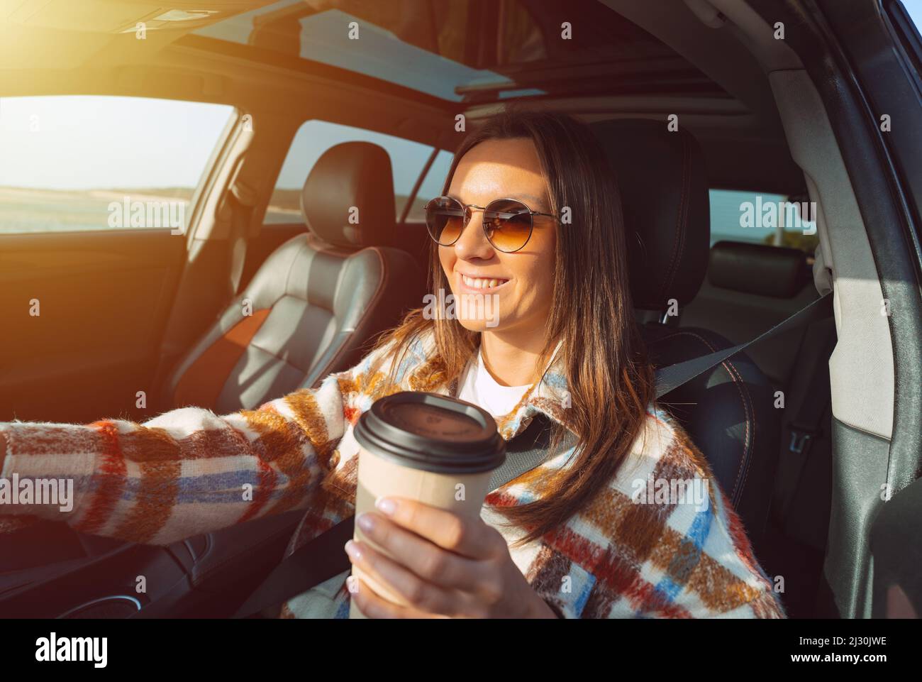 Felice sorridente bella donna che tiene la tazza di caffè e guida una macchina in una giornata di sole Foto Stock