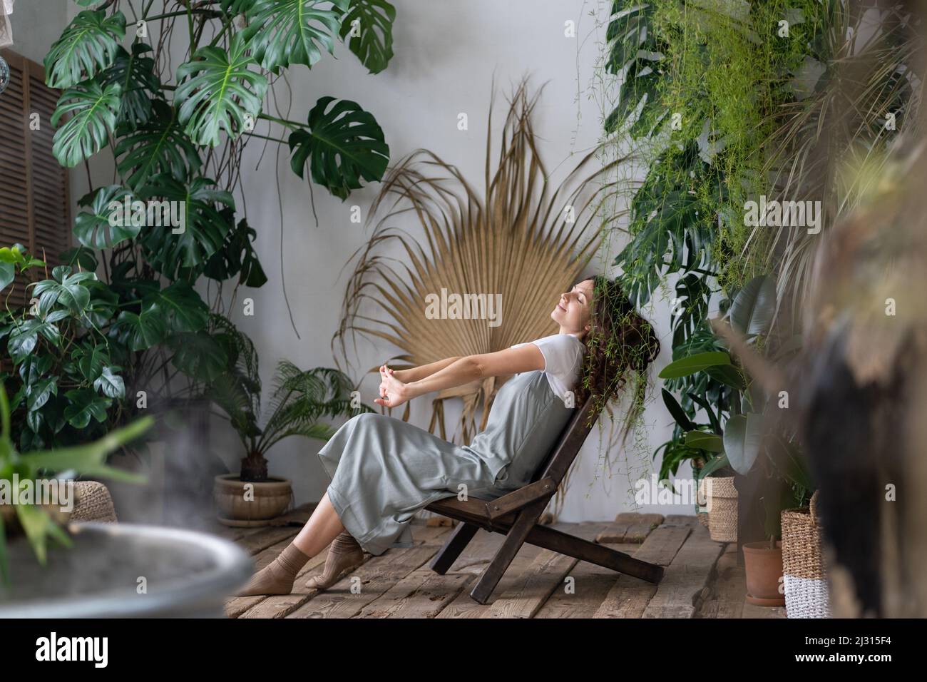 Donna sognante che si rilassa nel giardino tropicale in stile resort a casa, donna giardiniere riposante in serra Foto Stock