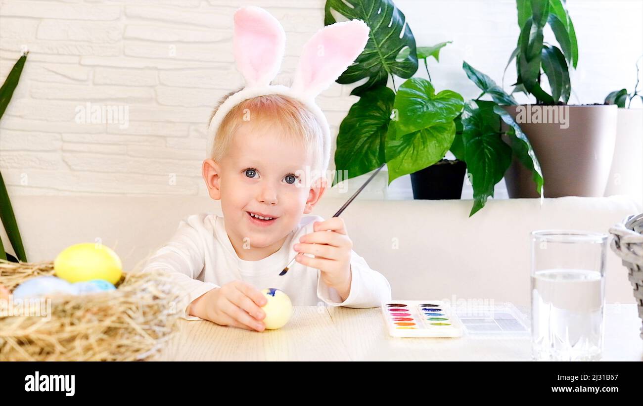 Un ragazzino in orecchie di coniglio dipinge entusiasticamente le uova di Pasqua con vernice. Concetto di vacanze religiose, fai da te, divertimento in famiglia, uovo di pasqua Foto Stock