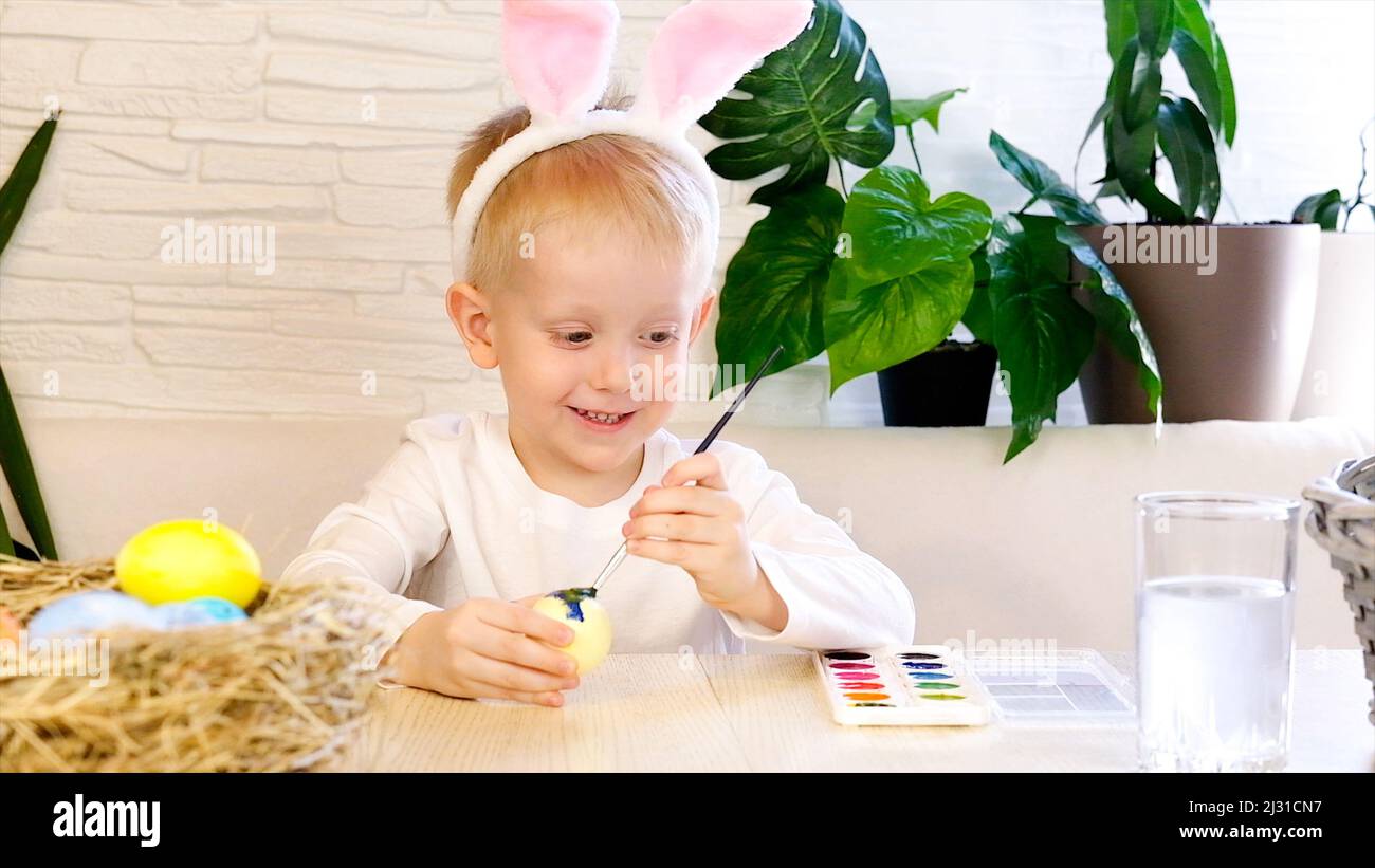 Un ragazzino in orecchie di coniglio dipinge entusiasticamente le uova di Pasqua con vernice. Concetto di vacanze religiose, fai da te, divertimento in famiglia, uovo di pasqua Foto Stock