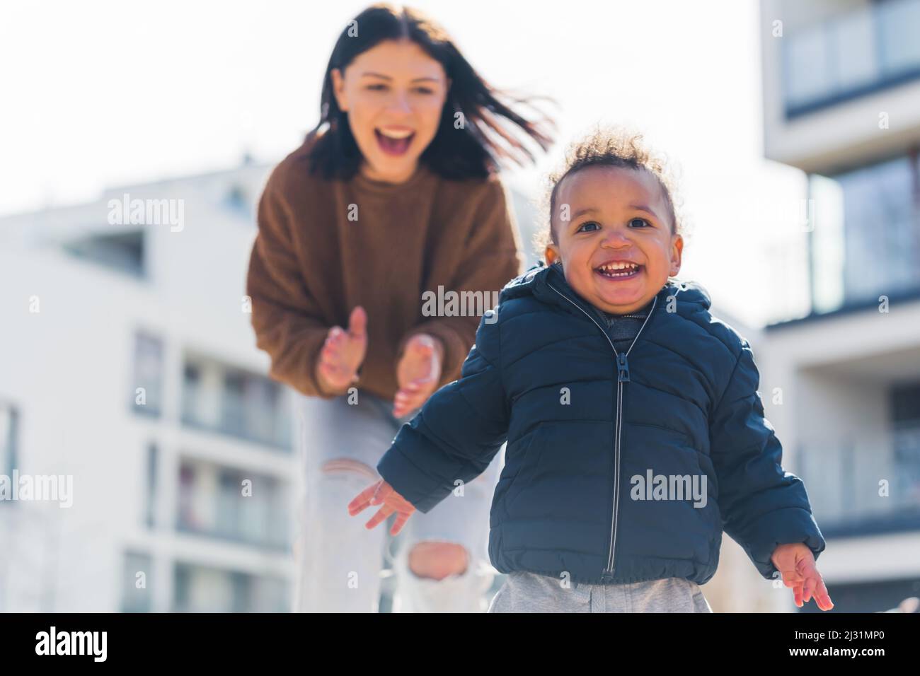 Ridendo bambino afroamericano con capelli ricci che corre davanti a sua madre che sta scattando sfondo urbano medio colpo. Foto di alta qualità Foto Stock