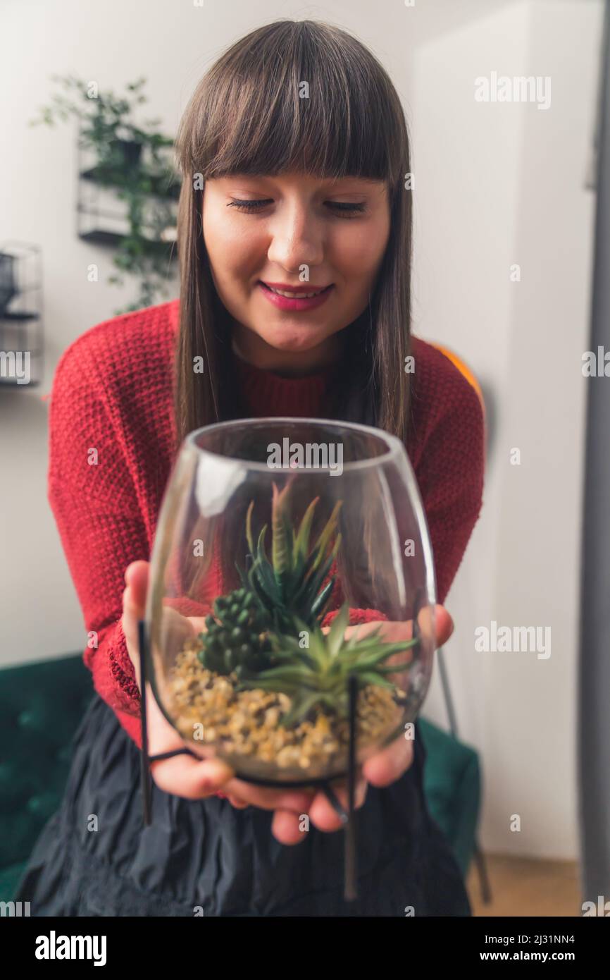 Colpo medio di graziosa giovane donna europea che tiene una pianta in una pentola di vetro, fuoco sul fronte casa sfondo . Foto di alta qualità Foto Stock