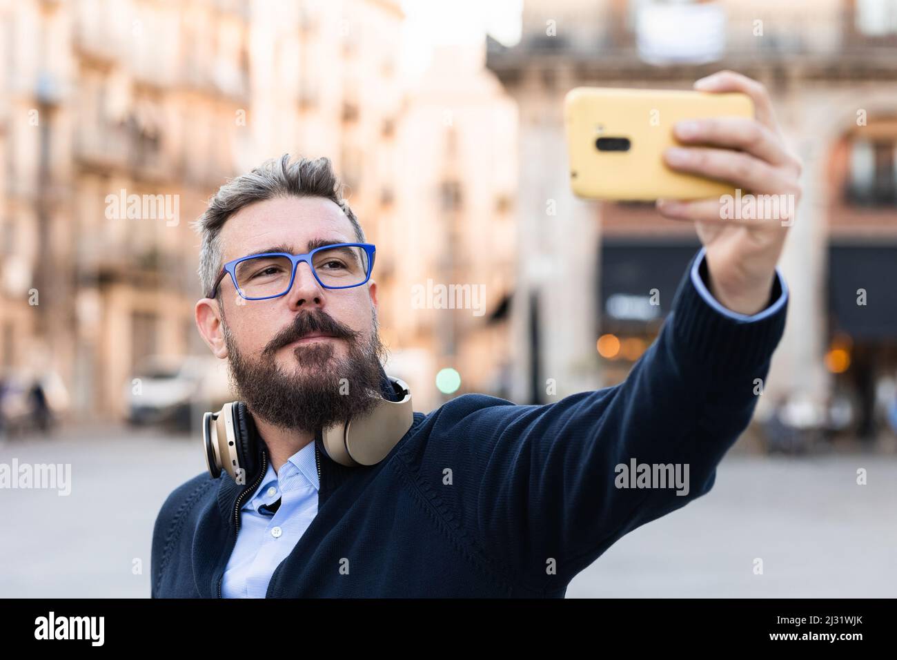 Uomo con occhiali da sole e cuffie senza fili con un selfie con un telefono cellulare all'aperto sulla strada. Foto Stock