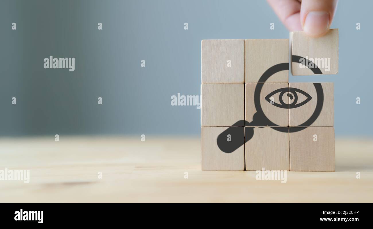 Ricerca, Ricerca, Ricerca della soluzione, analisi del concetto. La mano tiene i cubetti di legno con lente d'ingrandimento con i simboli degli occhi su sfondo grigio, copia s Foto Stock