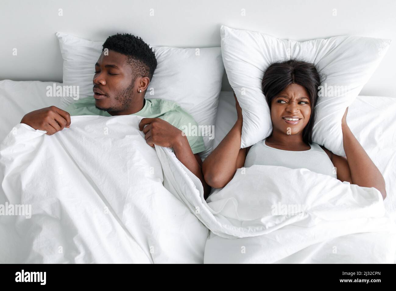 Infelice giovane afroamericana che copre le orecchie con cuscino, soffrendo di russare di marito addormentato Foto Stock