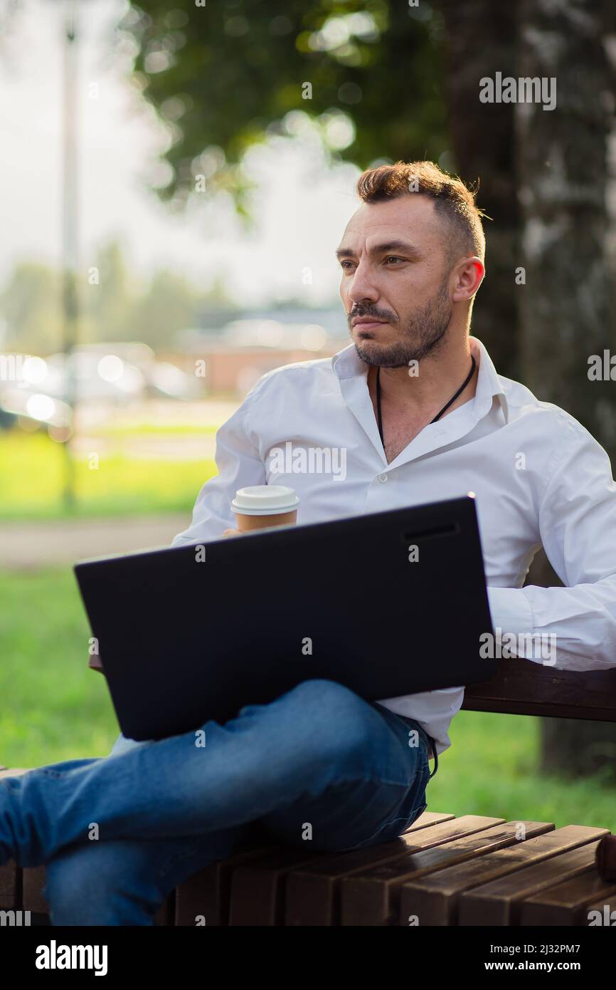 L'uomo sognante lavora nel parco con un computer portatile, beve caffè. Un giovane uomo su uno sfondo di alberi verdi, una calda giornata estiva di sole. Luce calda e soffusa, primo piano. Foto Stock