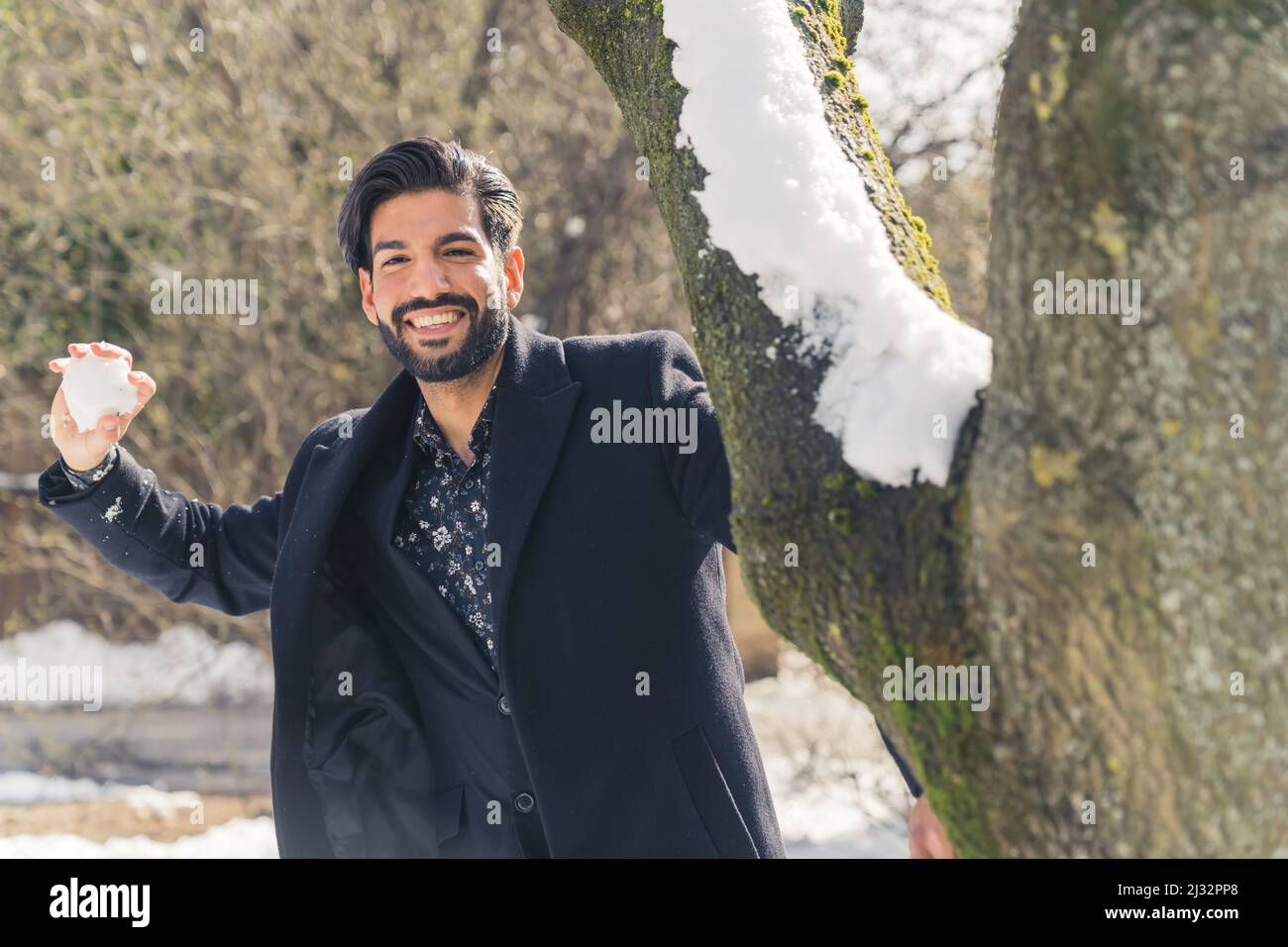 Allegro ed elegante uomo latino ispanico vestito con un cammello nero che lancia la palla di neve alla macchina fotografica - concetto di tempo libero invernale. Foto di alta qualità Foto Stock