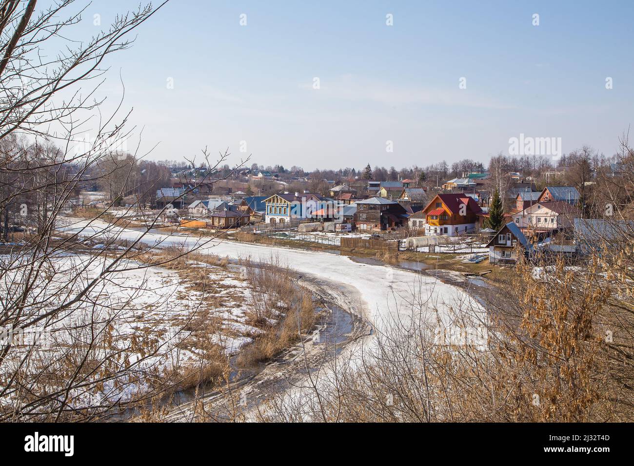 Un fiume curvo, ghiacciato che si estende in lontananza, un villaggio sulla riva. Primavera, neve si scioglie, pozzanghere e erba secca tutto intorno. Giorno, tempo nuvoloso, luce calda e soffusa. Foto Stock