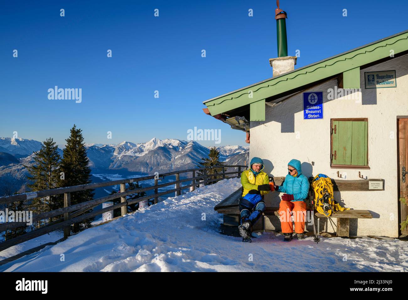 L'uomo e la donna che camminano con le racchette da neve sui loro zaini siedono sulla panchina davanti alla capanna coperta di neve e bevono il tè, Enzianhütte, Hochgern, Alpi di Chiemgau, alta Baviera, Baviera, Germania Foto Stock