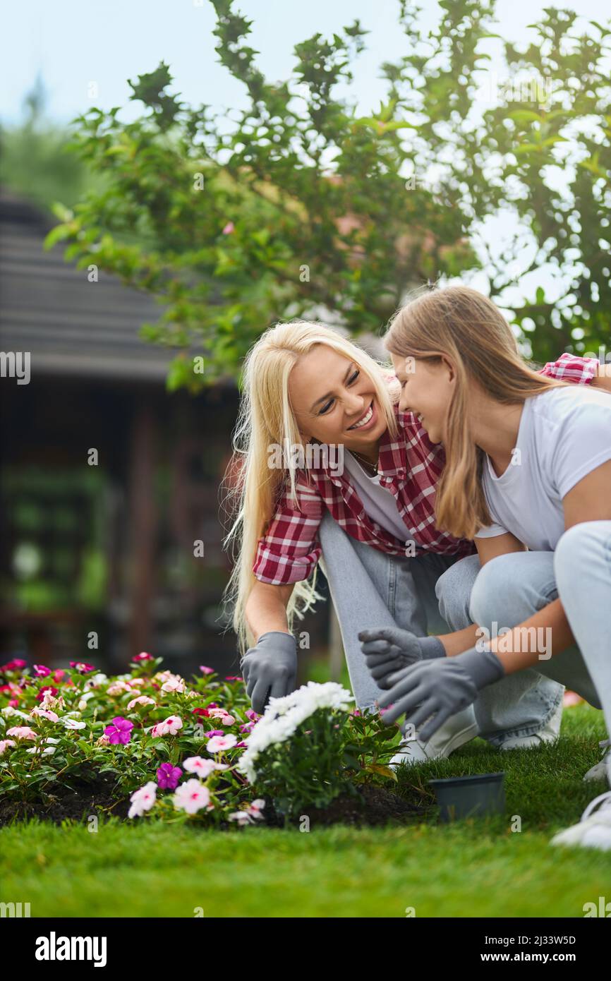 Cura della madre caucasica che insegna alla sua figlia teen di prendersi cura delle piante sul cortile. Donna e ragazza che squattano sul flowerbed, ridendo e abbracciando durante il processo di lavoro. Foto Stock