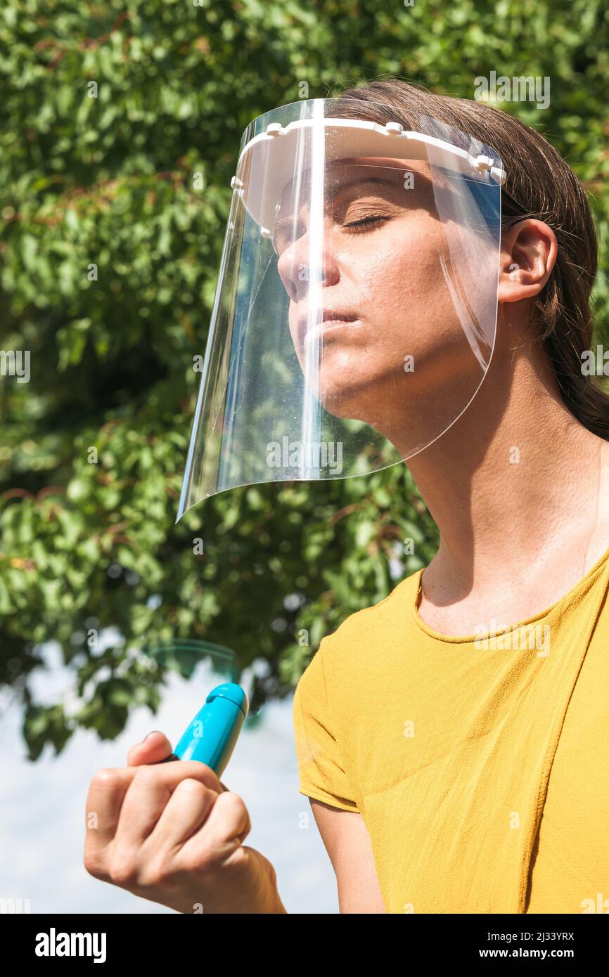 la giovane donna si raffredda con un ventilatore portatile indossando una visiera protettiva Foto Stock
