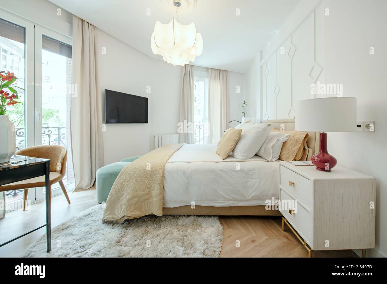 Camera da letto con un letto matrimoniale con cuscini di colore chiaro, comodini in legno chiaro e diversi balconi che si affacciano sulla strada Foto Stock