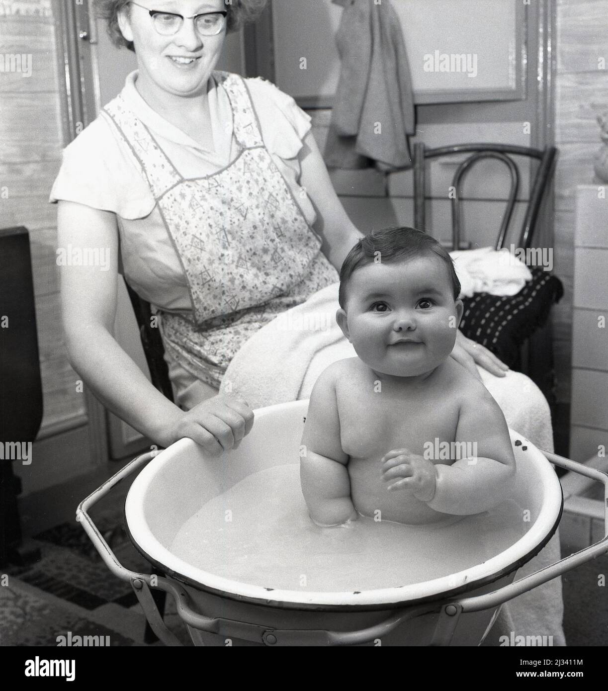 1959, storico, un bambino seduto in una ciotola di smalto in una cornice di metallo che ha un bagno, con la sua madre seduta accanto, portando un pinafore e con un asciugamano sul suo grembo, Stockport, Manchester, Inghilterra, Regno Unito. Foto Stock