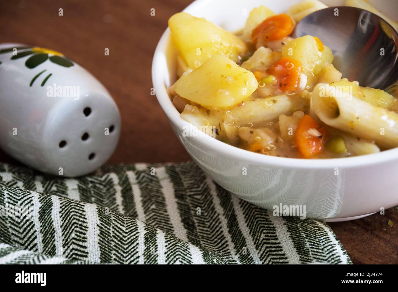 Ciotola di zuppa di verdure con patate, carote, quinoa, piselli e spaghetti, mostrando parte del cucchiaio all'interno della ciotola che si trova su un tavolo marrone con striscia verde Foto Stock