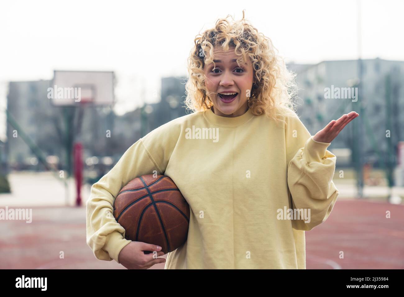 Stupito giovane bionda capelli ricci donna in un maglione giallo chiaro sul campo sportivo tenendo una palla di basket e mantenendo un'altra mano aperta ritratto copia spazio . Foto di alta qualità Foto Stock