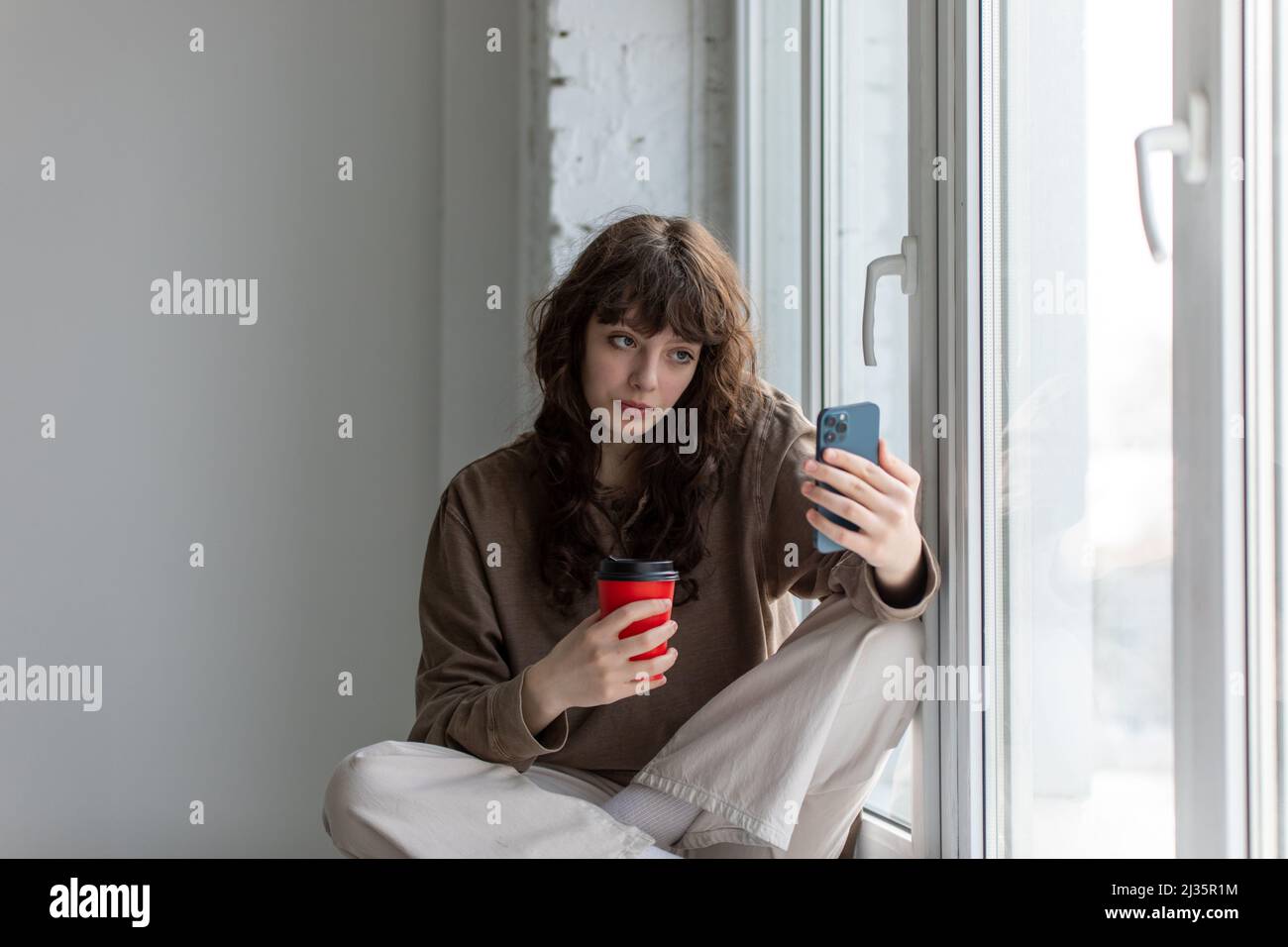Giovane donna attraente con una tazza di caffè nelle sue mani fa un selfie seduto sul davanzale. Foto Stock