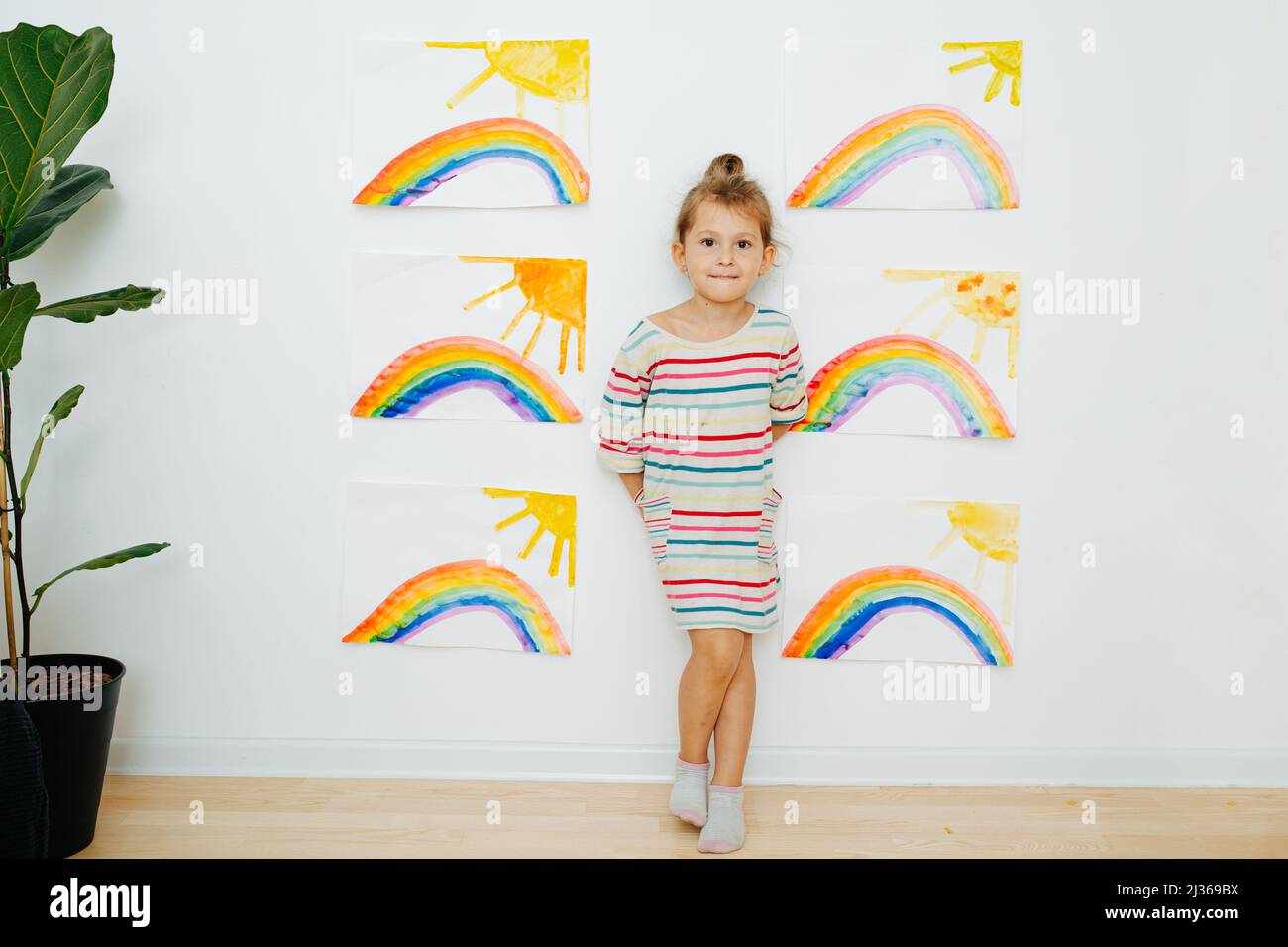 Piccola ragazza fiduciosa in una camicia vestito a righe accanto a sei dipinti simili di sole e arcobaleno ha dipinto. Posando di fronte alla parete. Foto Stock