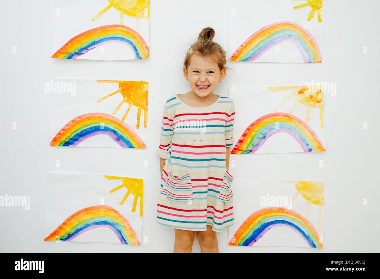 Ragazza energica allegra in una camicia vestito a righe accanto a sei dipinti simili di sole e arcobaleno che dipinse. Posando di fronte alla parete. Foto Stock