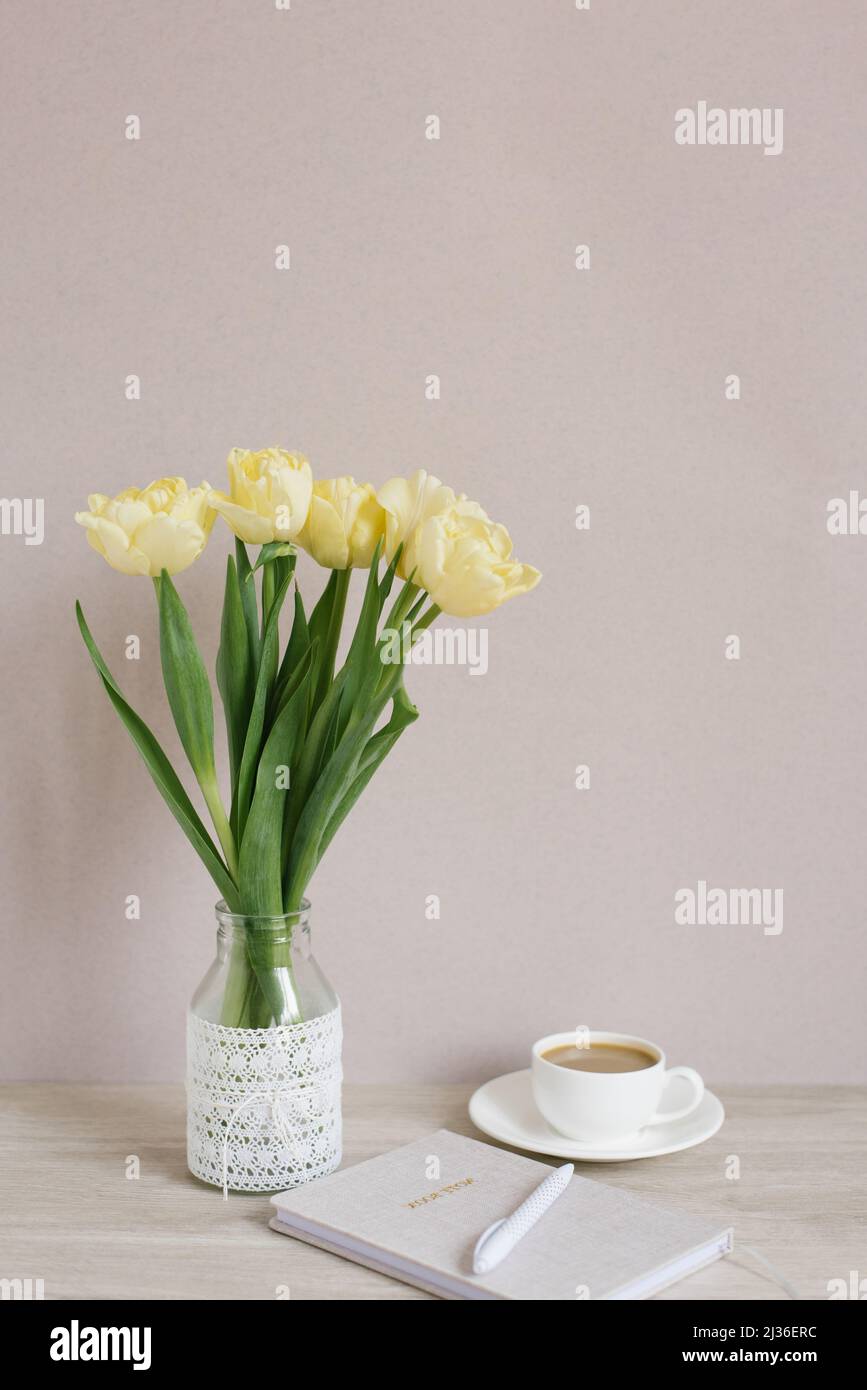 Tulipano giallo fiori in vaso sul tavolo, una tazza di caffè e un diario con una penna, parete grigio-beige come sfondo Foto Stock