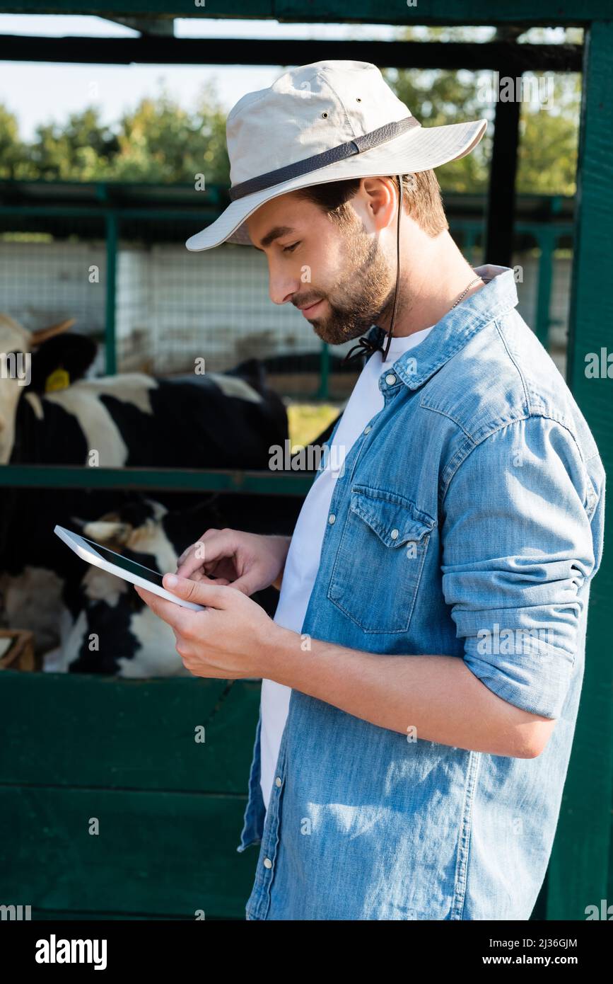 vista laterale di un agricoltore sorridente in cappello da sposa  utilizzando un tablet digitale vicino a un cowshed in azienda agricola Foto  stock - Alamy