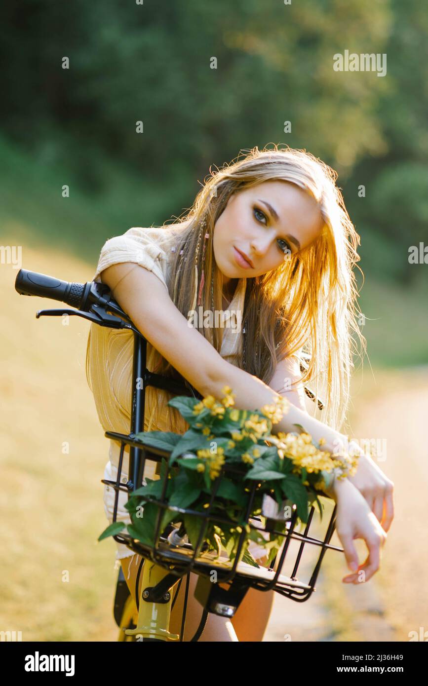 Una giovane bella donna tiene una bicicletta con fiori gialli di verbena in un cestino su una passeggiata all'aperto campagna Foto Stock