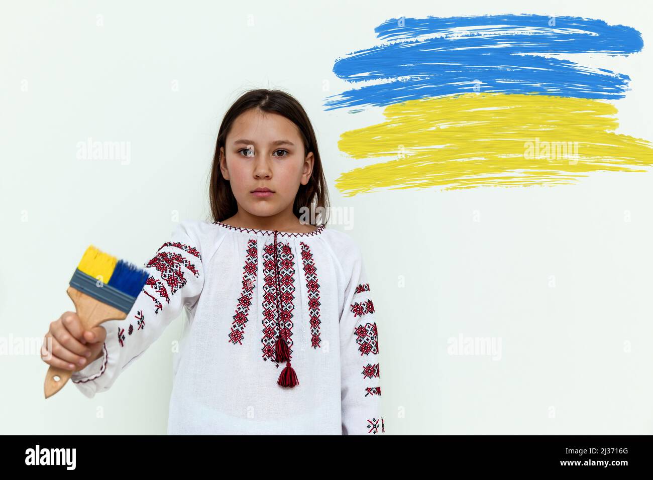 Una ragazza in un abito ricamato si erge contro una parete bianca, tenendo una spazzola dipinta di blu e giallo. La ragazza protesta contro la guerra. Ucraino-Russi Foto Stock