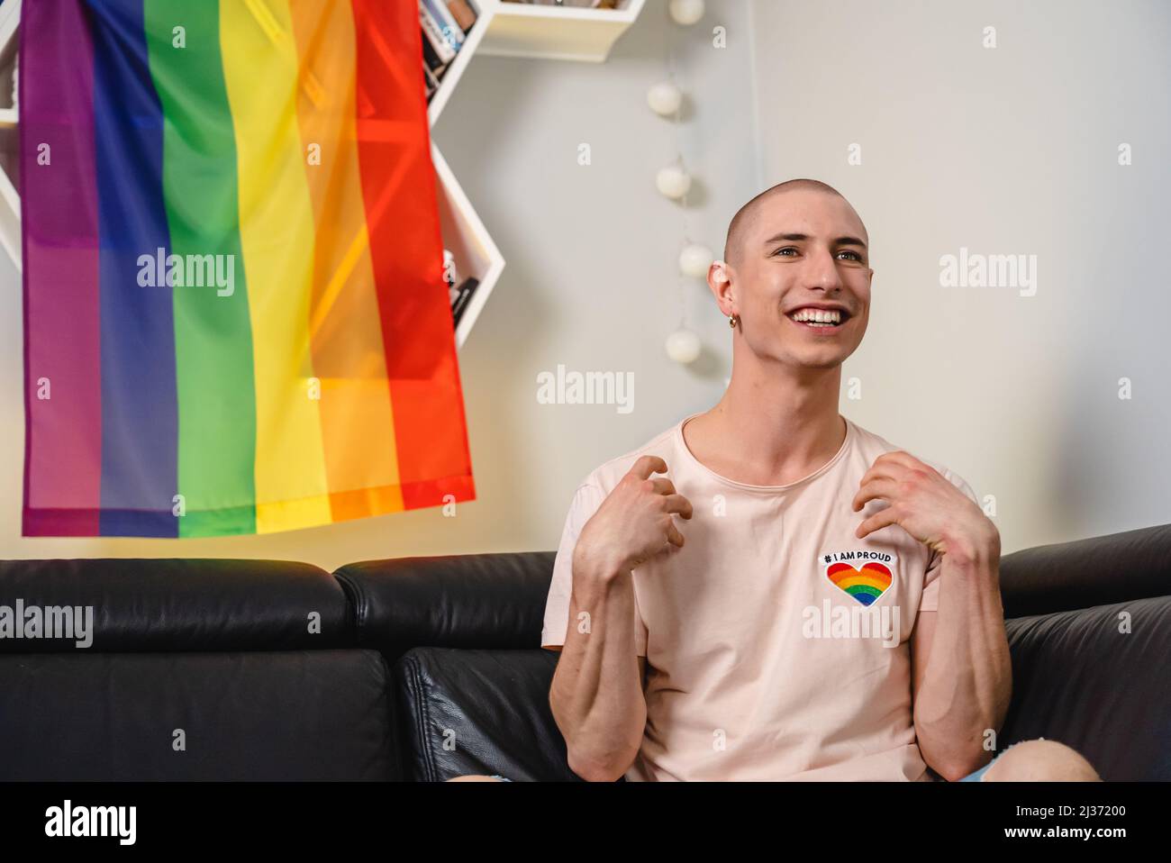 Calvo queer attivista caucasico seduto su un divano, guardando la tv, e sorridendo con le mani in su. Bandiera arcobaleno sullo sfondo. Foto di alta qualità Foto Stock