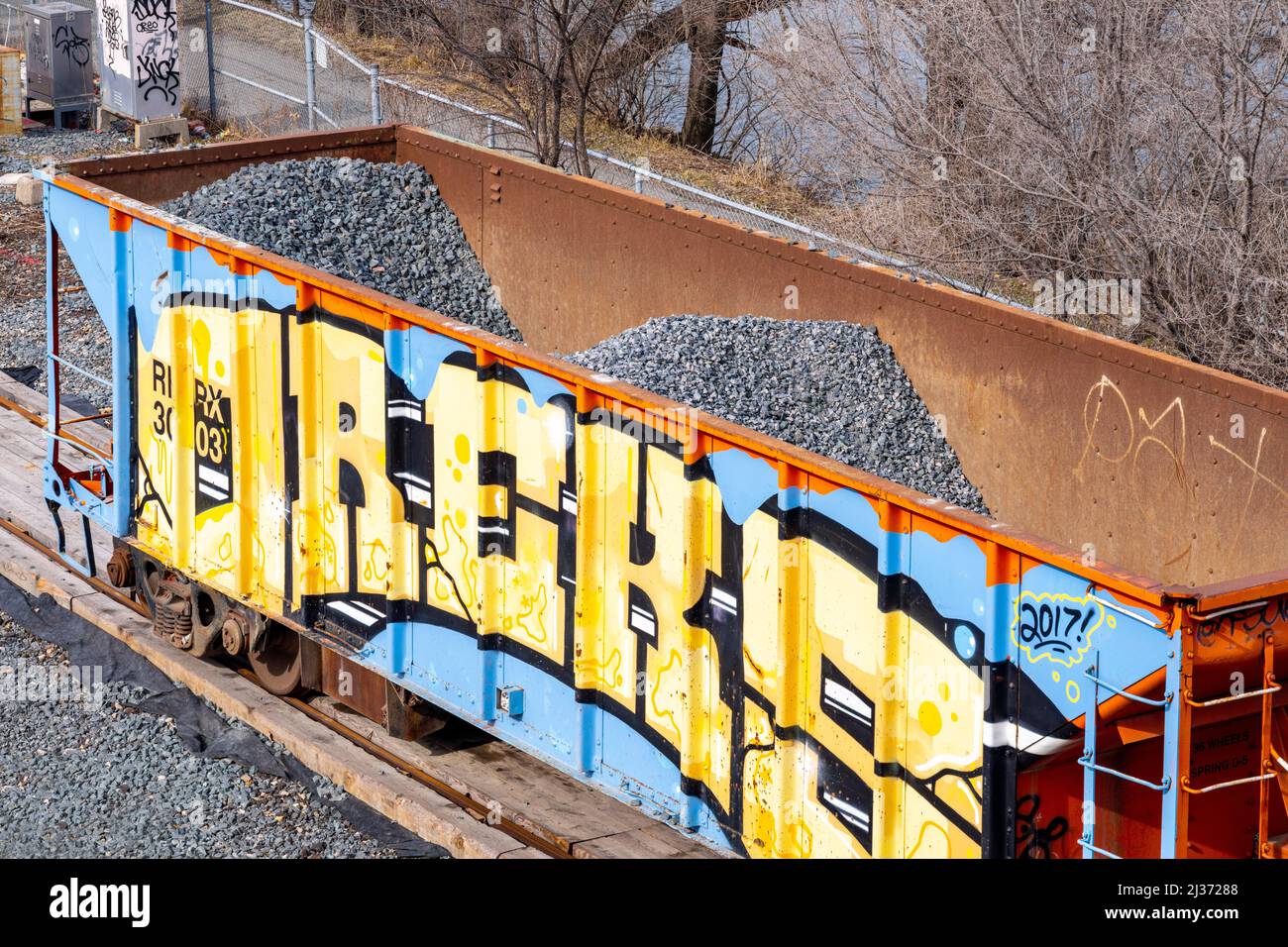 Un'arte di graffiti urbani è dipinta su un treno pieno di ghiaia. I materiali da costruzione e i veicoli sono fermi nel quartiere Queen St. East. Foto Stock