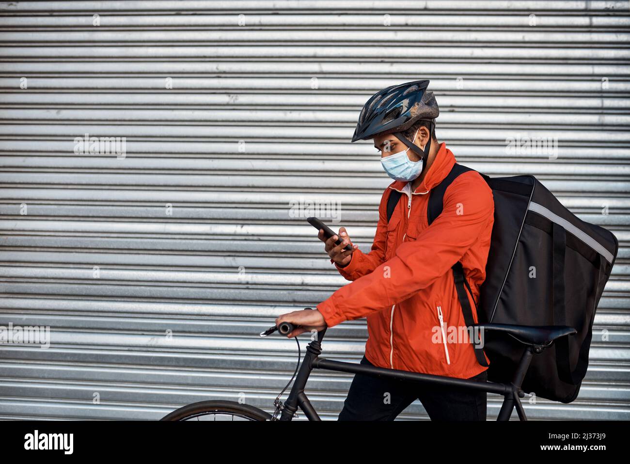 Verifica della posizione di consegna successiva della sua app. Colpo di un uomo mascherato usando il suo cellulare mentre fuori sulla sua bicicletta per una consegna. Foto Stock