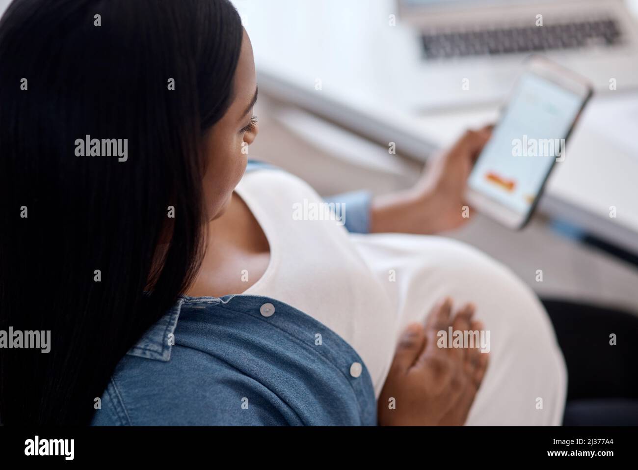 Questa app mi ha aiutato a rendere questa permanenza memorabile. Scatto di una donna incinta usando il suo cellulare mentre si siede a casa. Foto Stock