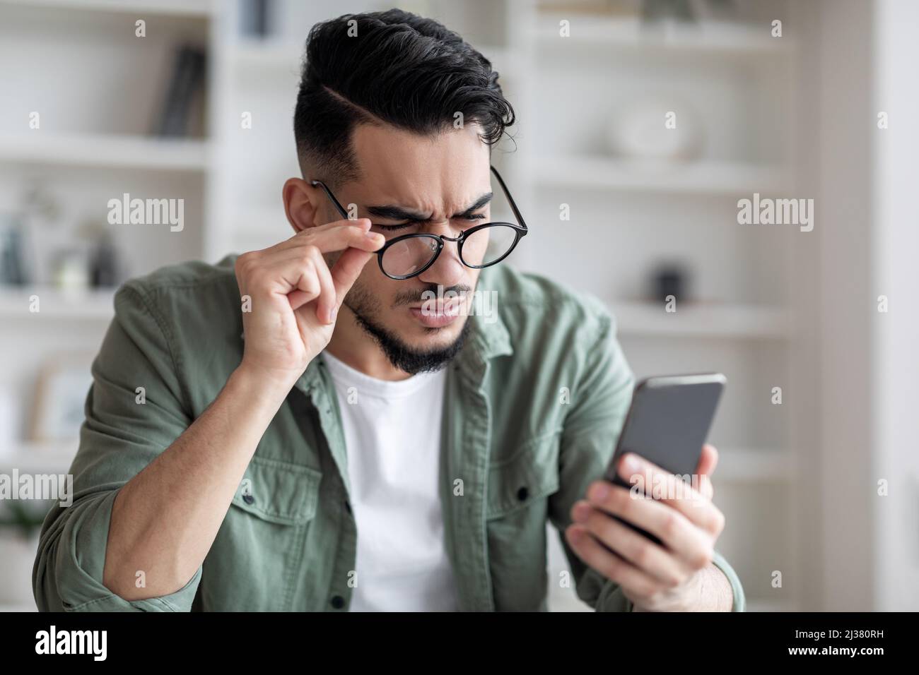 Problemi di vista concetto. Giovane uomo arabo in occhiali guardando lo schermo dello smartphone Foto Stock