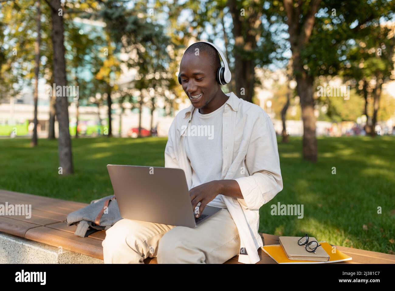 Ragazzo nero sorridente che si rilassa con un computer portatile e le cuffie nel parco dopo le lezioni, godendo di trascorrere il tempo all'aperto Foto Stock