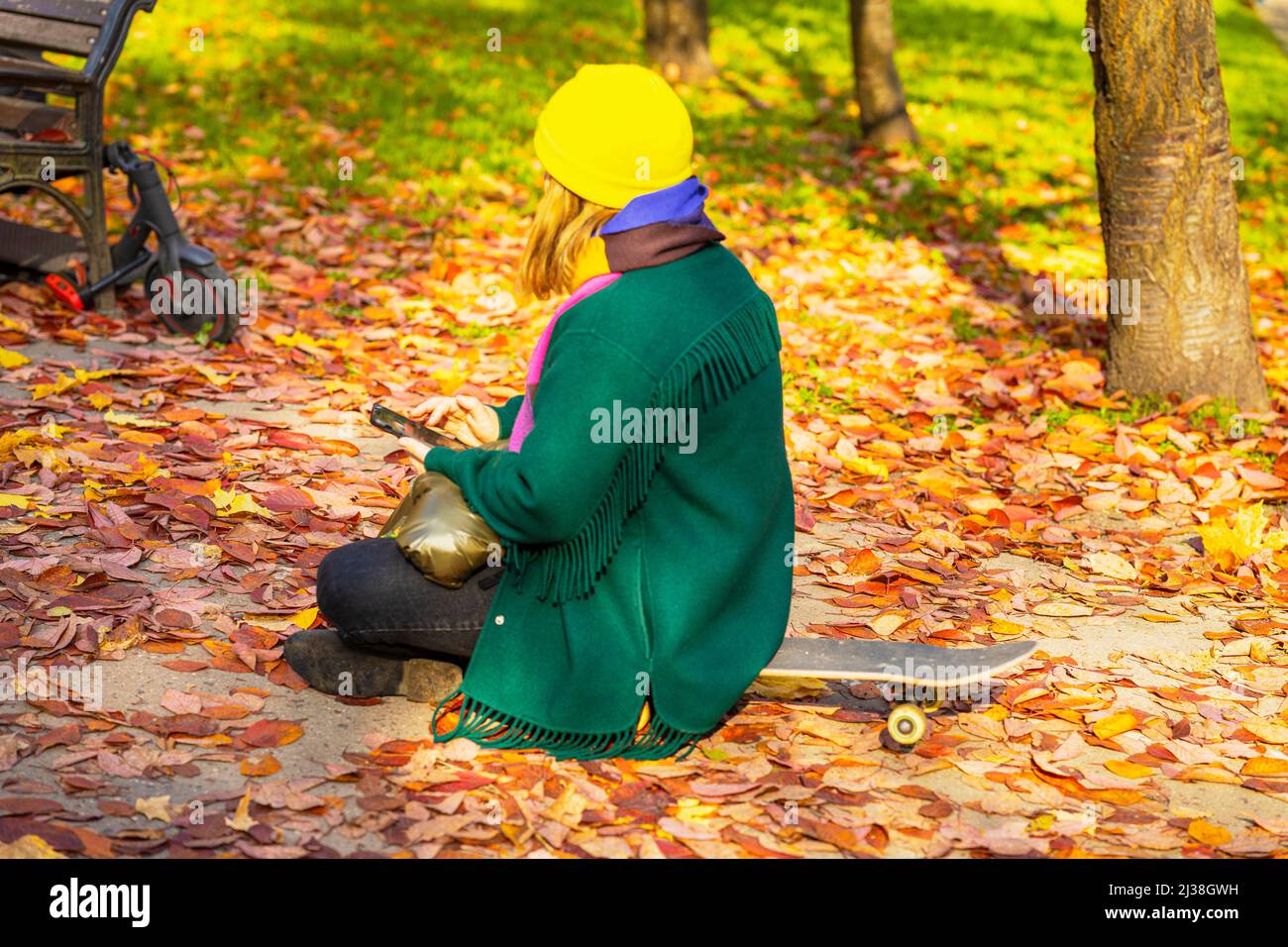 Giovane ragazza irriconoscibile che usa il telefono in un parco coperto in autunno foglie mentre si siede sullo skateboard. Autunno giorno di sole Foto Stock