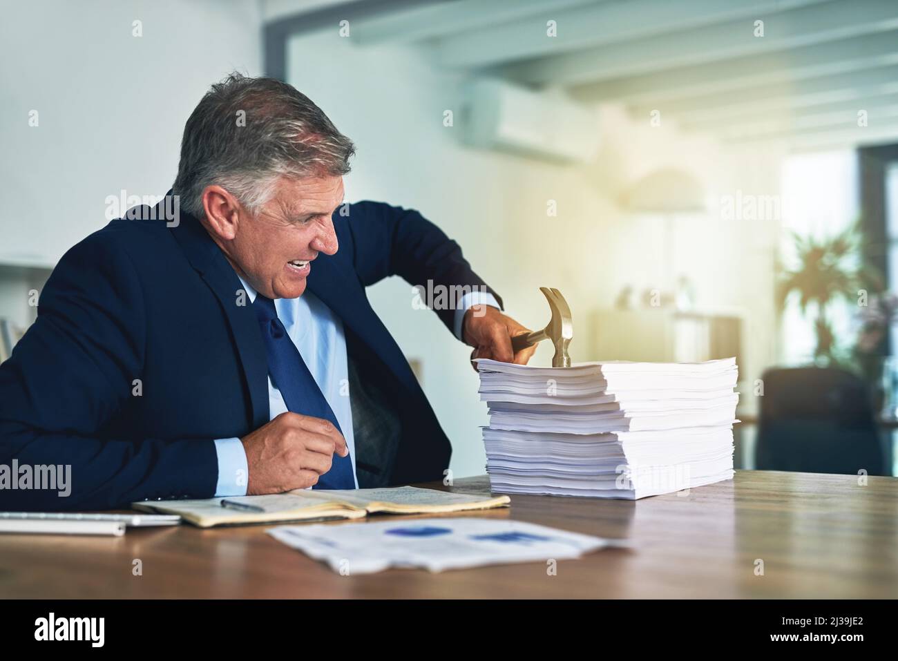 Ha un approccio alternativo al lavoro cartaceo. Colpo di un uomo d'affari frustrato colpendo un mucchio di lavoro di carta con un martello. Foto Stock