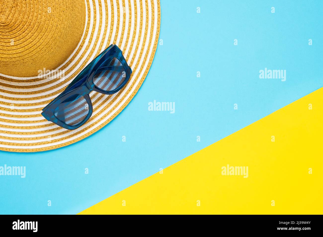 Concetto di vacanza estiva. Accessori da spiaggia, cappello a righe e occhiali da sole su sfondo giallo-blu. Vista dall'alto, spazio di copia Foto Stock